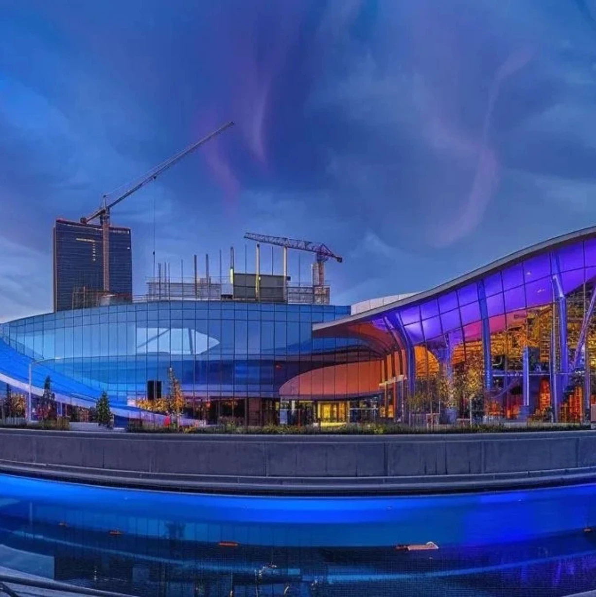 Modern architecture with blue lighting, reflecting in water. Cranes in the background.