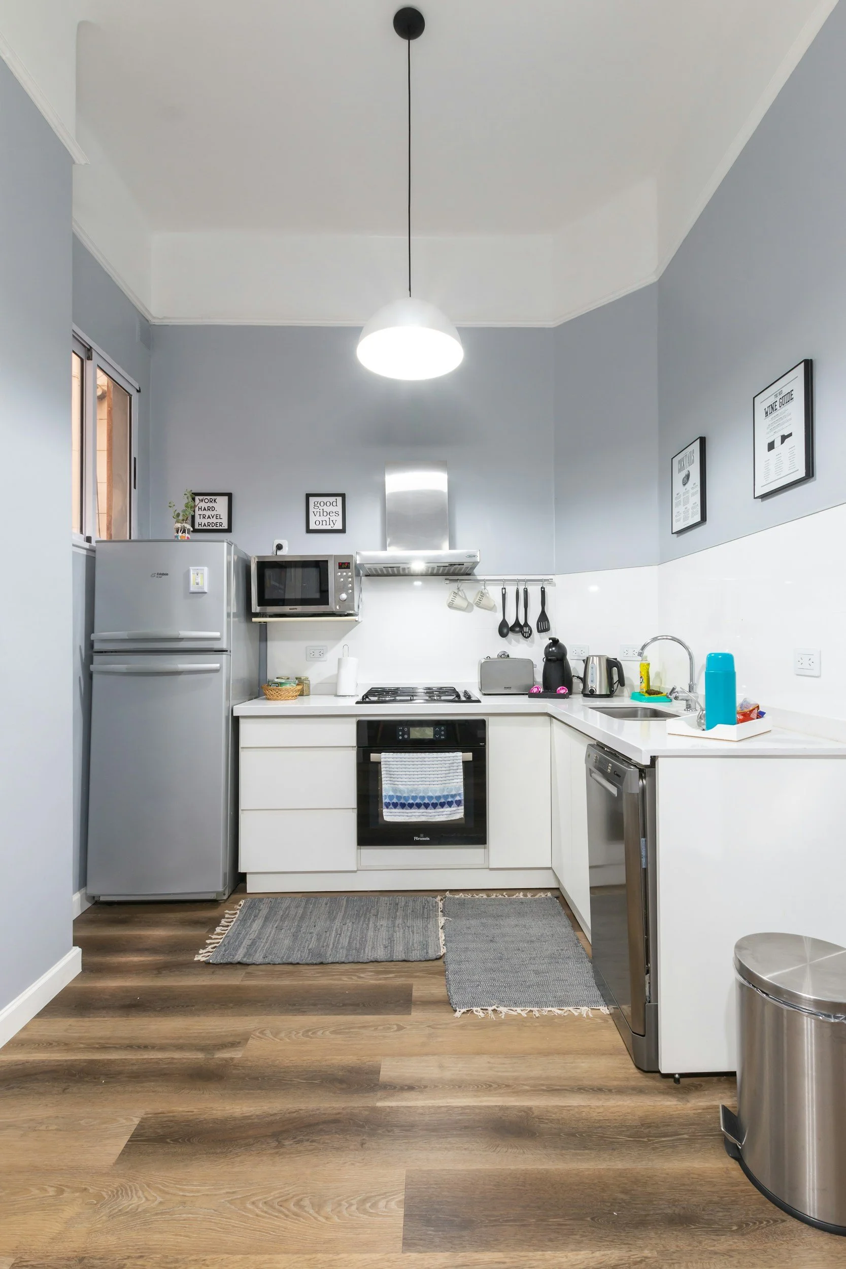 Modern kitchen with stainless steel appliances, white cabinets, and wood flooring. "Good vibes only" art on the wall.